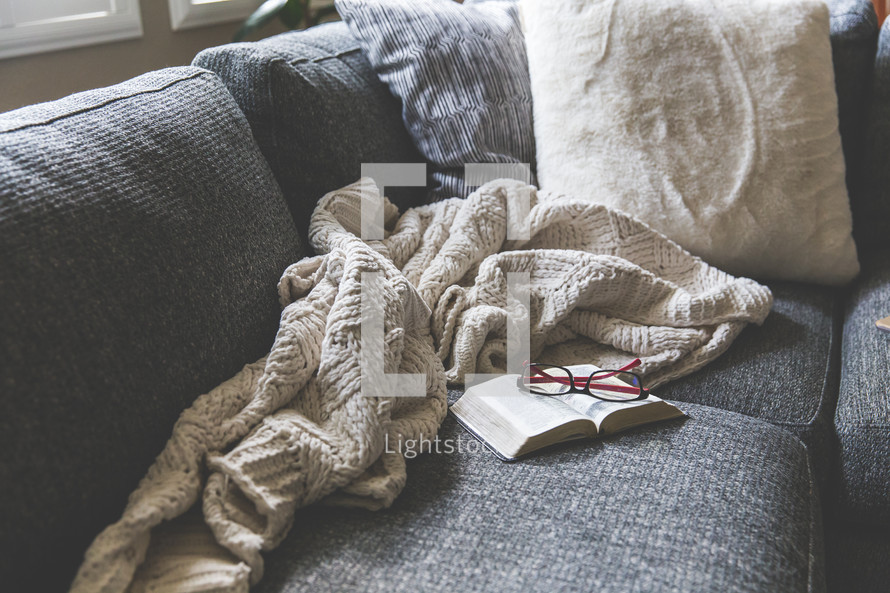 reading glasses, blanket, and open Bible on a couch 