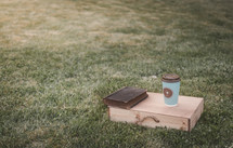coffee cup with cross, wood box, and Bible on grass 