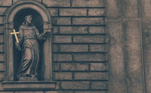 Statue of a saint holding a gold cross  in an alcove of a brick wall.