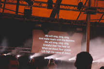 lyrics on a projection screen and worship leaders singing on stage 