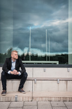 a business man sitting on steps with a reflection of a stormy sky in a window 