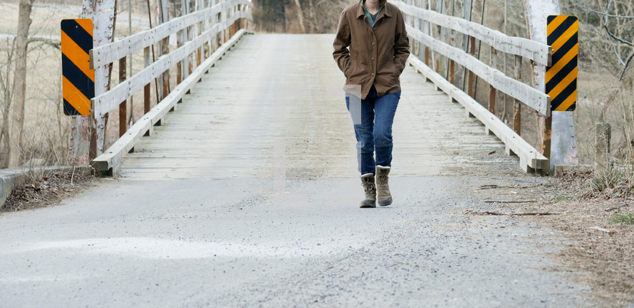 a woman in boots and a coat walking across a wooden footbridge 