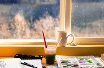coffee mug and paint supplies in front of a window 