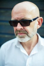 A bald man in a white shirt with dark sunglasses.