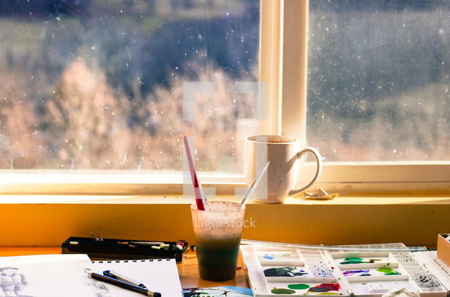 coffee mug and paint supplies in front of a window 