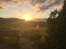 Sun setting behind hills and shining on a wooden bench in the countryside.