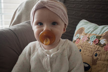 an infant girl with a pacifier in her mouth sitting on a couch 