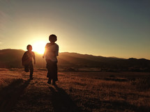 Silhouette of young boys running through the grass by the mountains at sunset.