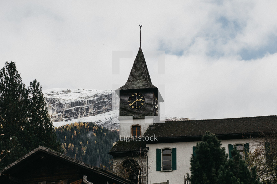 clock on a steeple and snow capped mountain peaks in the clouds 