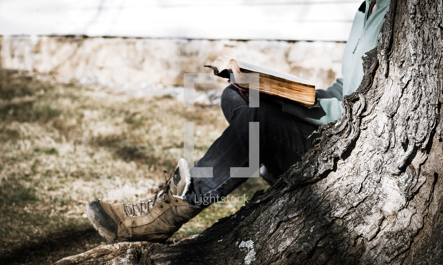 a woman in boots reading a Bible under a tree 