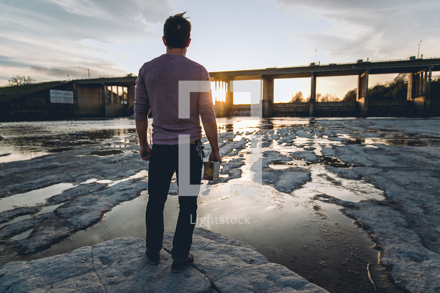 A man standing on a rocky shore looking out at the sun setting behind a bridge holding a Bible at his side 