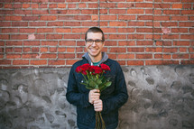 A man standing in front of a brick wall holding red roses. 