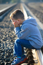 A little boy sits on a railroad track with his face in his hands.