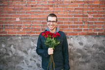 A man holding red roses in front of a brick wall. 