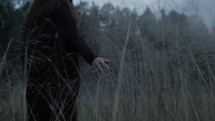 a woman walking in a field of tall grasses touching them with her hand 