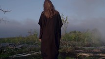 a woman in a flowing cloak walking through a misty mountaintop landscape 