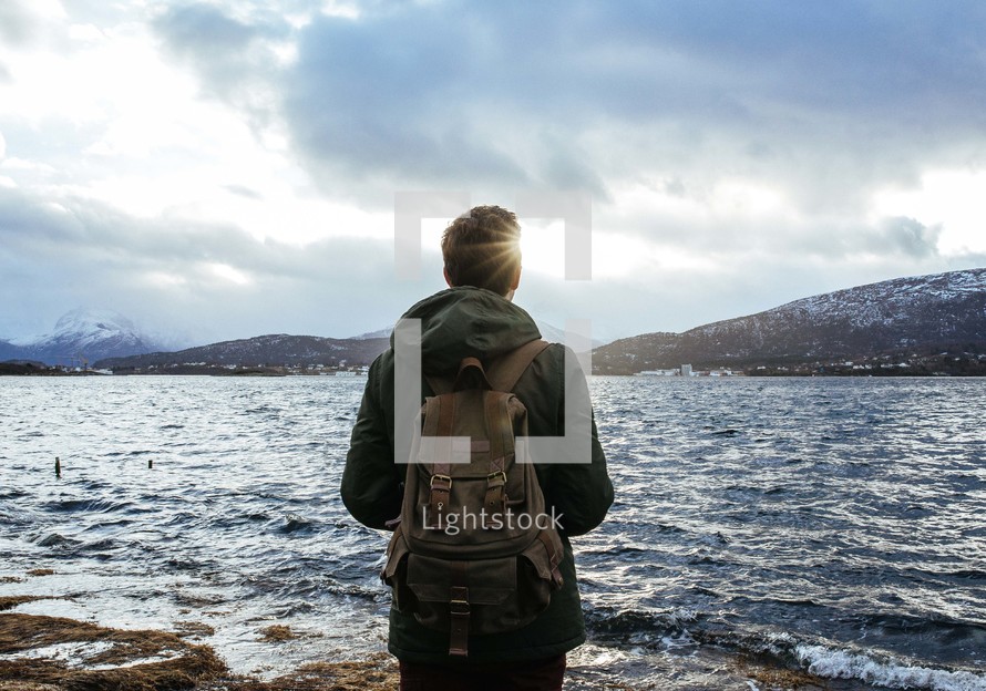 man with a backpack standing on a shore looking out at the water 