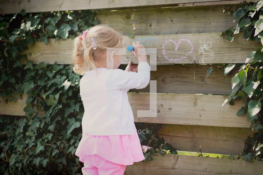 a girl child coloring a fence with chalk 