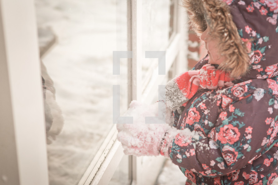 A little girl in winter clothes looks at her reflection in a window.