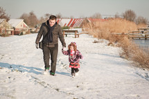 A man and a little girl hold hands and run through the snow.