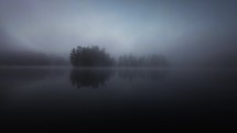 A drone footage fly over toward a small lake island covered in pine tree forest the lake is misty and fog has settled in on the cool fall day