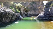 Aerial shot drone flies under pedestrian bridge with woman on it and over kayaker on river below as it passes under car bridge then an ancient bridge opening into large green valley