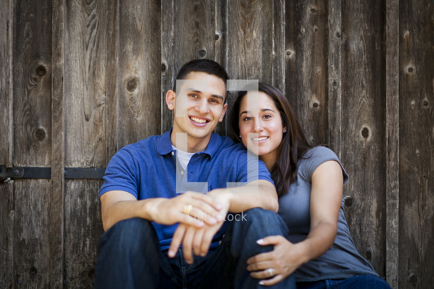 couple sitting on the ground together in front of a wood fence