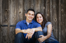 couple sitting on the ground together in front of a wood fence