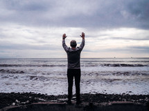 man standing with his hands raised in front of the ocean 