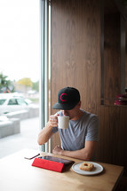 a man sitting in front of an iPad drinking coffee at a window seat 