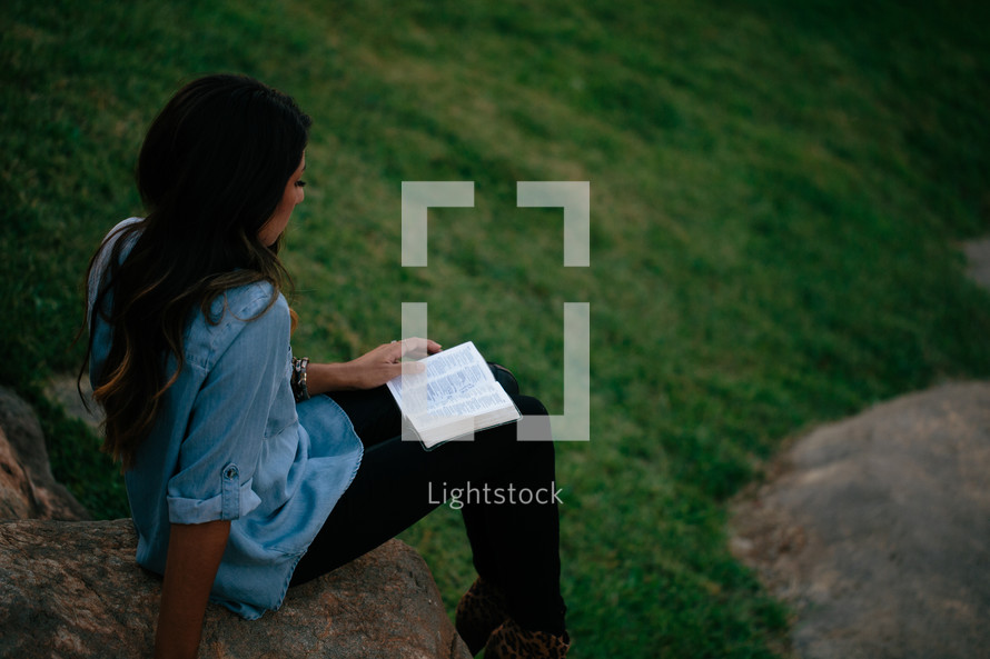 A young woman sits outdoors on a rock reading a Bible.