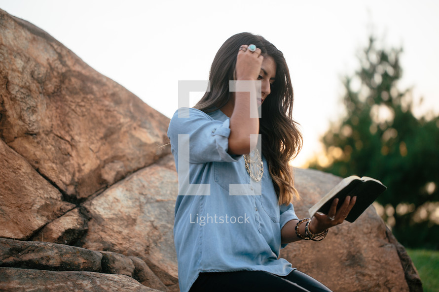 a woman reading a Bible sitting on a rock putting her hair behind her ear 
