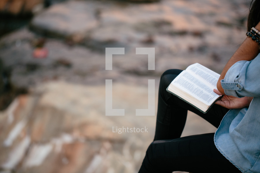 A young woman sits outside on a rock and reads the Bible.