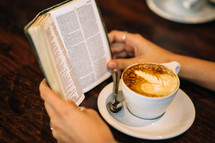 Hands on a wooden table with an open Bible and a cup of coffee.