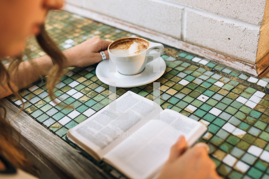 Woman reading a bible on a mosaic tile table with a cup of coffee.