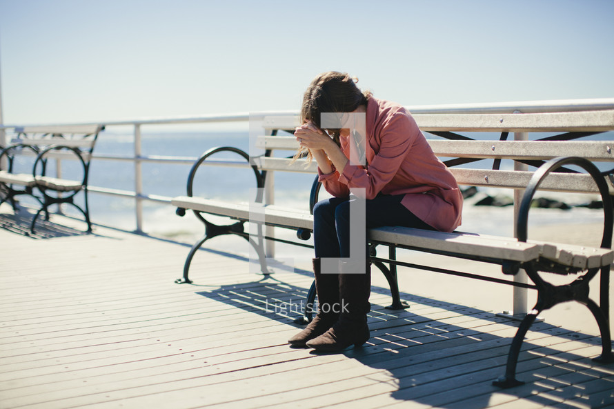 Woman sitting on bench on ocean side deck with elbows on her knees and her head resting on her folded hands.
