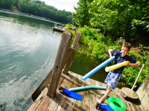a boy child with pool noodles on a lake dock 