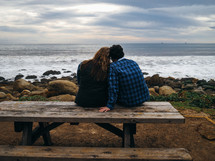 Couple sitting close together on a picnic table looking out at the ocean.