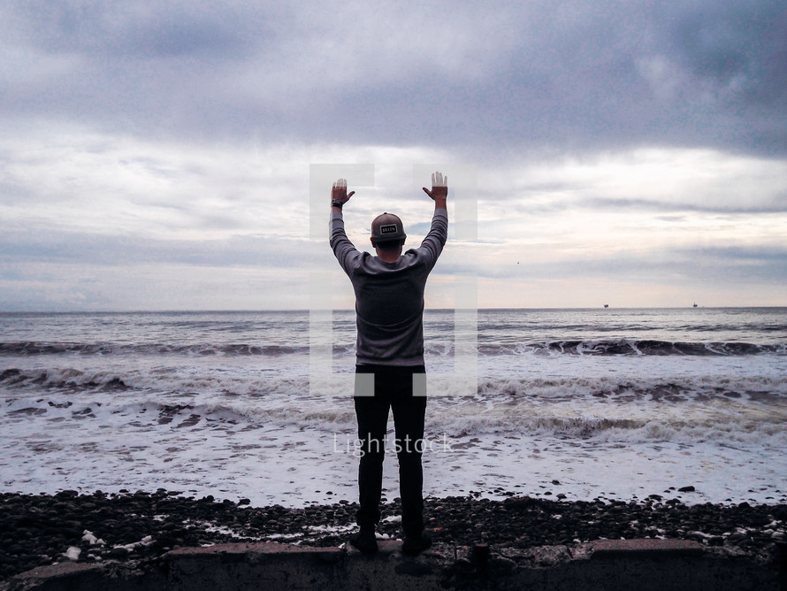 man standing with his hands raised in front of the ocean 