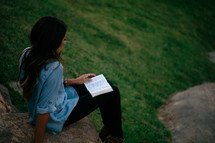 A young woman sits outdoors on a rock reading a Bible.