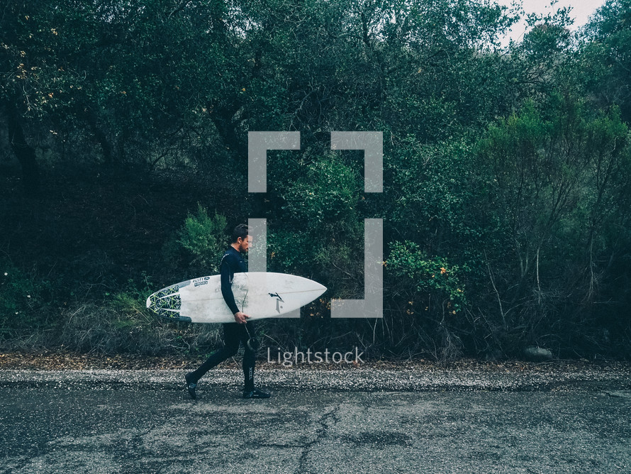 Man in a wetsuit carrying a surfboard along an asphalt road.