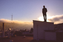 A silhouetted man stands a top a building a dawn