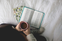 a woman reading a Bible sitting on her bed drinking coffee 