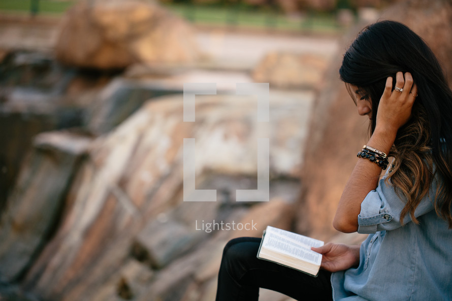 a woman sitting on a rock reading a Bible outdoors 