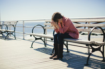 Woman sitting on bench on ocean side deck with elbows on her knees and her head resting on her folded hands.
