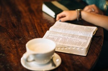 Hands om a wooden table with an open Bible and a cup of coffee.