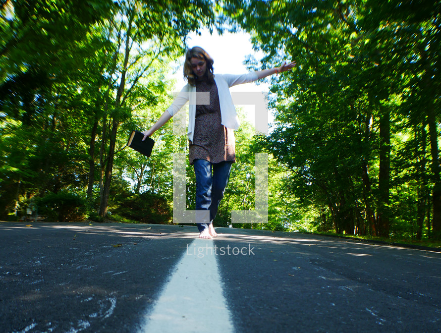 a teen girl walking on the center lines of a road holding a Bible 