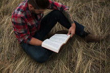 man in a plaid shirt sitting on the ground reading a Bible 