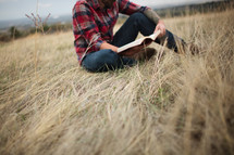 A man in a plaid shirt reading a Bible sitting on the ground 