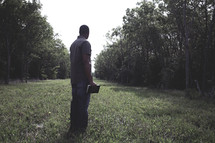 Man standing in a tree-lined, grassy field holding a Bible.